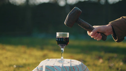 a hand holding a mallet about to smash a glass of red wine on a small table