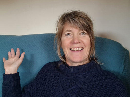 Close up of Jo wearing a navy blue woollen polo neck jumper against a teal blue sofa and smiling at the camera. One hand is raised in a little wave.