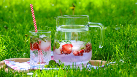 close up of a jug and glass of water, decorated with fresh strawberries, sitting on sunlit green grass