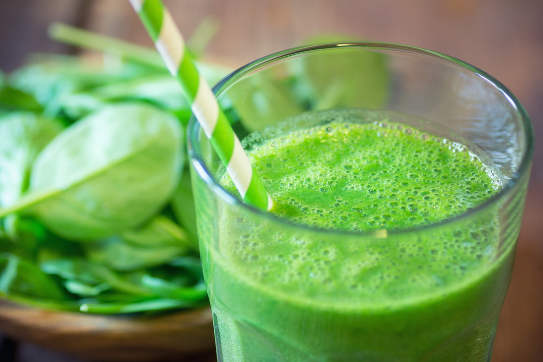 green smoothie in a glass with a green and white striped straw and a bowl full of spinach leaves sitting behind the glass on a wooden table