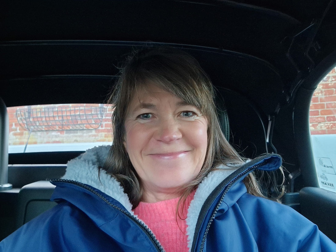 Close up headshot of Jo smiling at the camera, sitting behind the wheel of the car and wearing a blue coat over a pink jumper