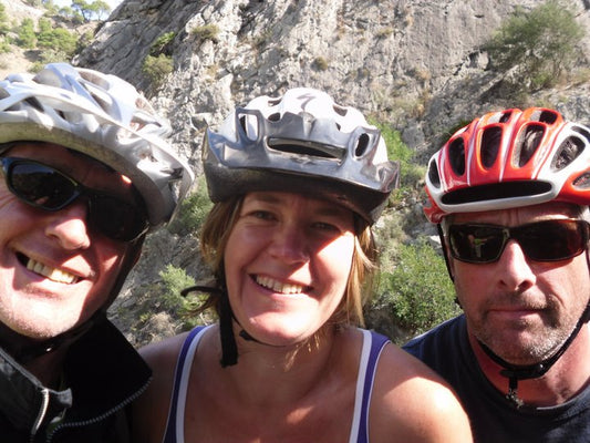 Selfie of Jo, Stuart and friend with cycle helmets on, smiling into the camera. Background grey rocky mountain with some greenery.